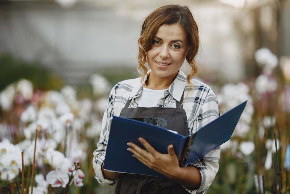 A woman stands outdoors in a lush garden with blooming white orchid flowers, holding an open blue gardening checklist folder. She is dressed casually in a plaid shirt layered over a white top and wears a black apron, suggesting she is engaged in garden maintenance or landscaping work. The garden features a level lawn area with trimmed grass, garden beds bordered with soil and plants, and a gravel pathway visible behind her. In the background, there are mature trees and blurred foliage, indicating a well-maintained, natural garden environment. The lighting is soft and natural, highlighting the textures of the plants, the wet soil, and the woman's friendly expression, reflecting ongoing garden care activities aligned with the Teddington riverside garden care checklist for gardeners coordinated by gardenersrichmonduponthames.org.uk.